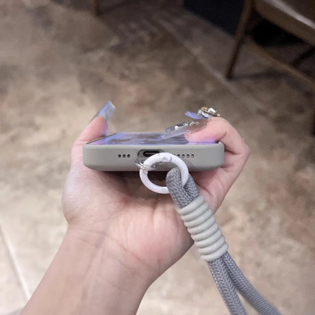 A hand holding a smartphone in a protective case with a gray braided lanyard attached.