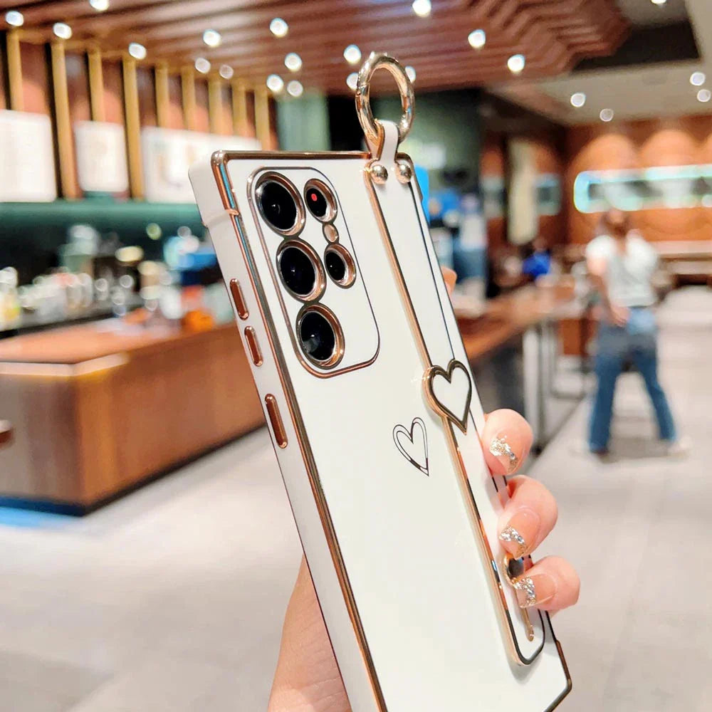 White smartphone case with gold edges, a heart-shaped stand, and a finger strap.