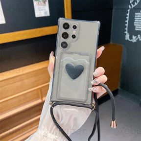 A clear phone case with a black heart-shaped pop socket and a black lanyard strap.