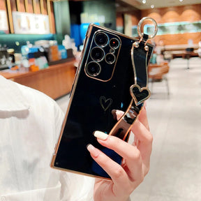 Black smartphone with a gold-trimmed case, a heart-shaped design, and a black and gold heart-shaped wrist strap.