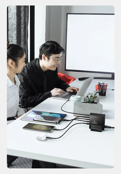 Laptop computer being used by two people at a desk with multiple electronic devices connected.