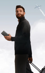 Man in a black outfit holding a smartphone and luggage, with an airplane visible in the sky behind him.