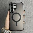 A person holding a dark, translucent phone case with a black MagSafe ring and an elongated ’I’ shape below it, featuring a prominent camera cutout for multiple lenses and corner bumper protection.