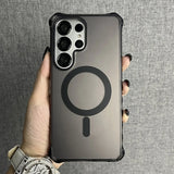 A person holding a dark, translucent phone case with a black MagSafe ring and an elongated ’I’ shape below it, featuring a prominent camera cutout for multiple lenses and corner bumper protection.