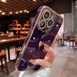 A person holding a purple smartphone with a gold-trimmed case, decorative studs, and a heart-shaped charm.