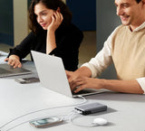 Two people working on laptops at a desk with various electronic devices visible.