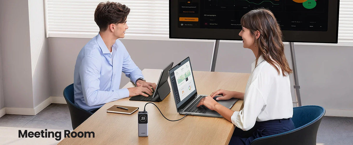 Two people sitting at a table with laptops in what appears to be a meeting room setting.