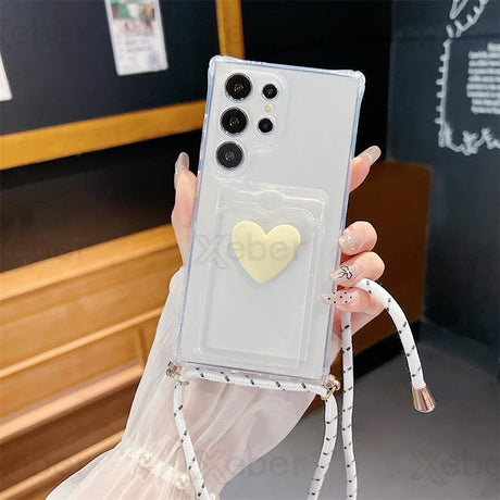 White smartphone in a clear case with a heart-shaped pop socket and a crossbody strap.