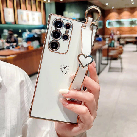 White smartphone in a rose gold-trimmed case with a heart-shaped wrist strap and a small heart drawing.