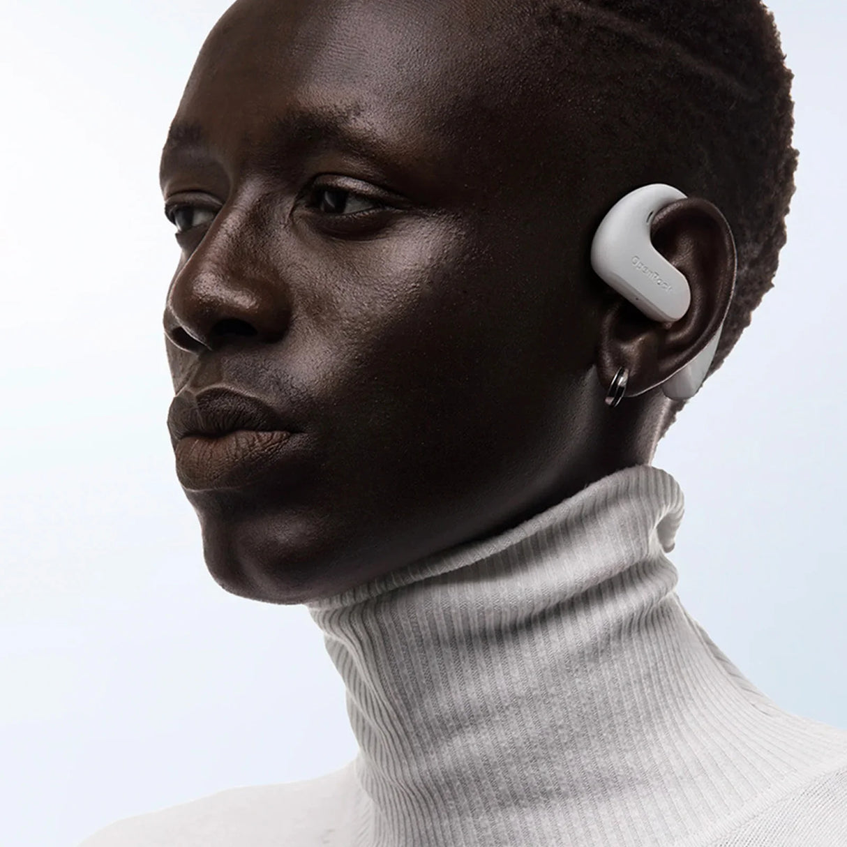 Wireless earbud worn by a person with dark skin in a white turtleneck sweater.